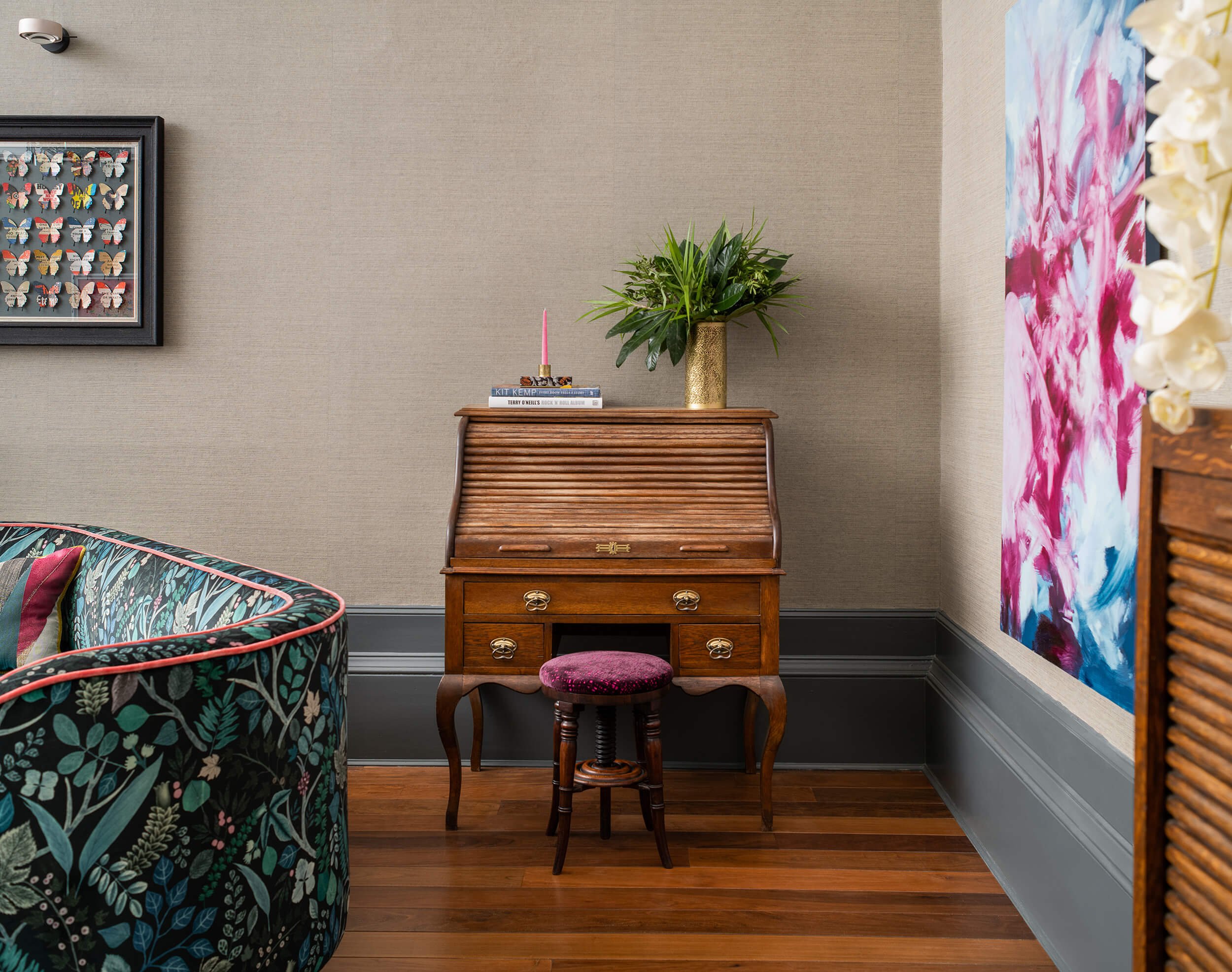 Eclectic room featuring antique roll-top desk, floral sofa, framed butterfly art, and vibrant pink & blue abstract painting.
