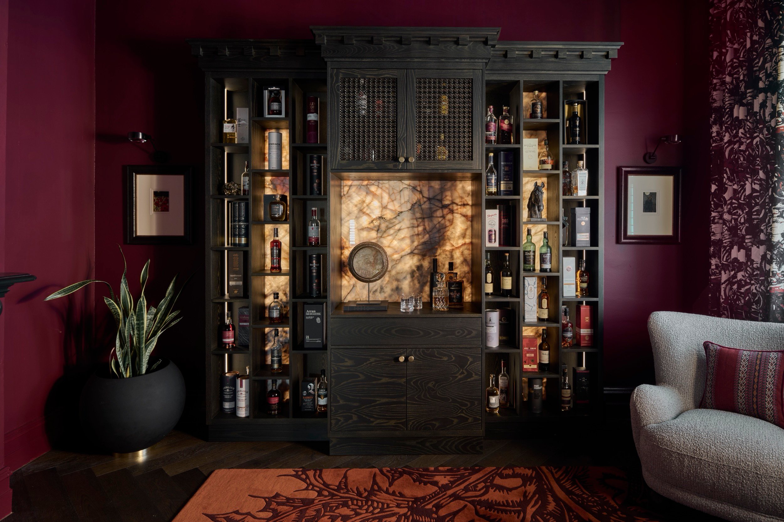 Dark wood built-in bar with backlit onyx, spirits collection, deep burgundy walls, textured armchair, and ornate rug.
