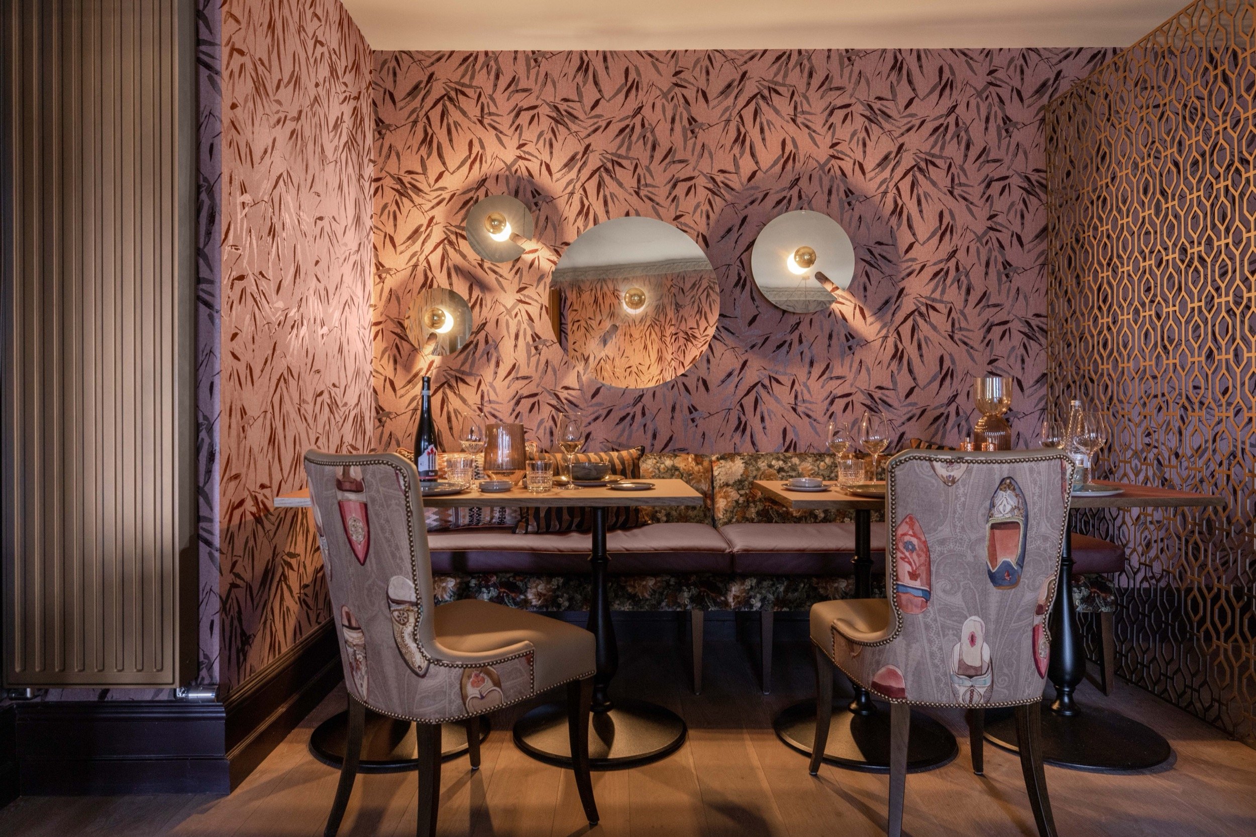Intimate dining room with blush bamboo wallpaper, eclectic chairs, plush booth seating, modern wall sconces, and a gold geometric divider.