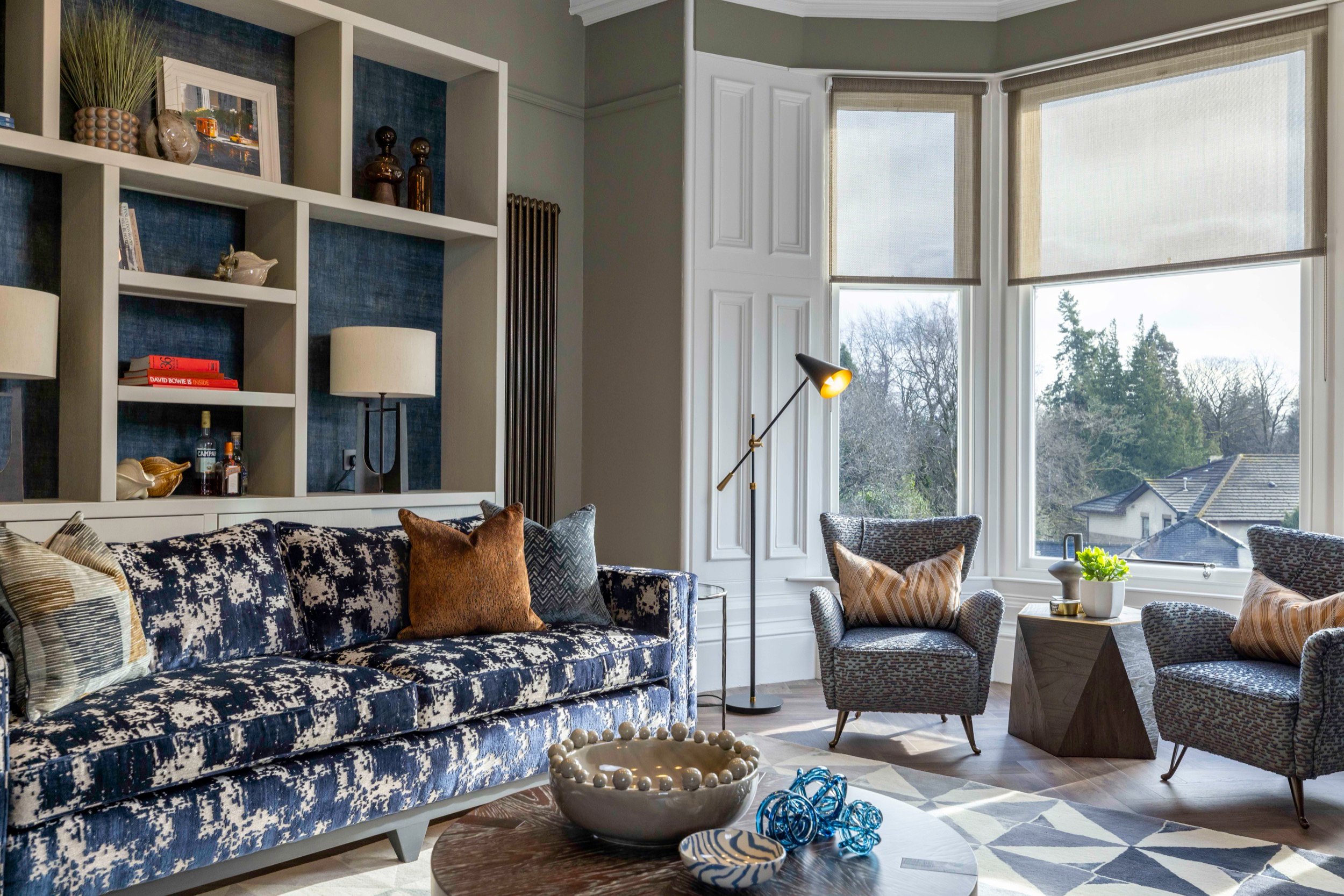Stylish lounge with blue abstract sofa, built-in shelves, two armchairs, patterned rug, and bright bay window view.