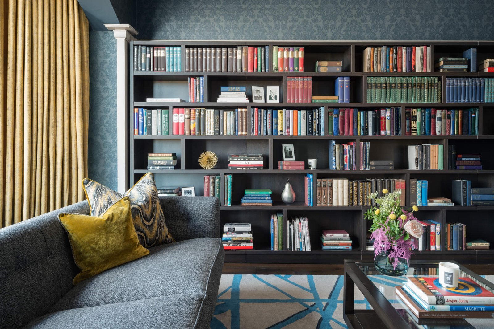Sophisticated living room with a dark floor-to-ceiling bookshelf filled with books, grey sofa, yellow pillows, and gold curtains.