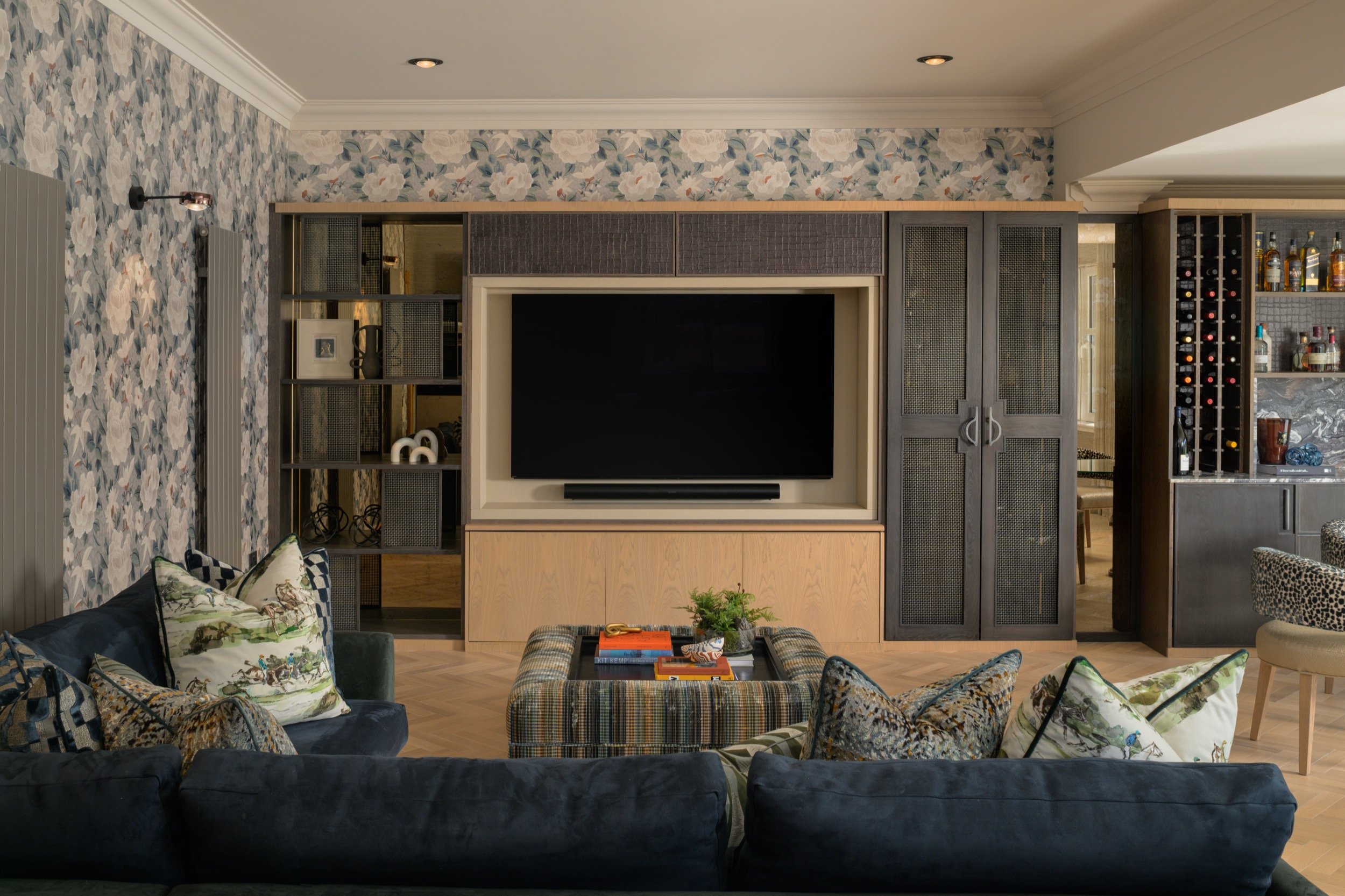 Elegant living room with floral wallpaper, built-in media unit, dark blue velvet sofa, plaid ottoman, and wet bar.