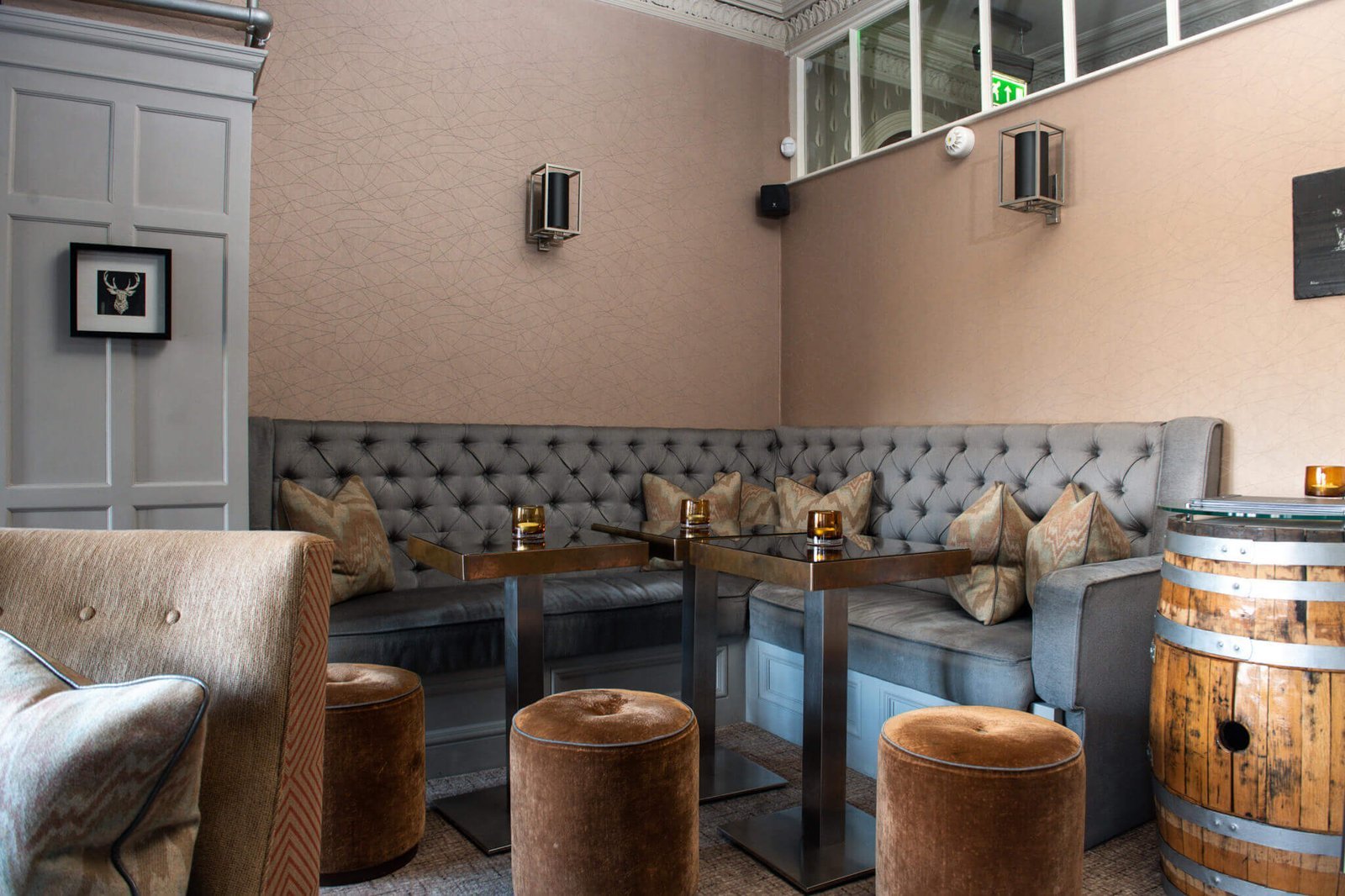 Elegant lounge bar with grey tufted banquette, sleek square tables, velvet stools, geometric wallpaper, and a whiskey barrel.